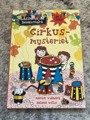 Cirkusmysteriet - LasseMajas Detektivbyrå - Färgglad barnbok med hårda pärmar från serien LasseMajas Detektivbyrå. Boken heter 'Cirkusmysteriet' och är skriven av Martin Widmark och illustrerad av Helena Willis. Omslaget har cirkustema med illustrationer i gult, rött, blått och svart.