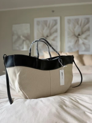 Otroligt fin slutsåld stor praktisk canvas tote bag med läderdetaljer i naturvit/svart färg - Stor tote bag i kraftig canvas i naturvit med svarta detaljer i läderimitation. Fyra handtag i olika längder, magnetknäppning, fodrad insida och dekorativa sömmar. Perfekt som vardags-, jobb- eller shoppingväska.  Mått: 28 × 54 × 15 cm.