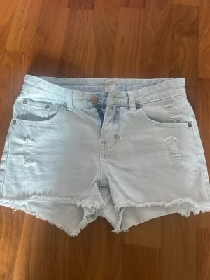 Ljusblåa jeansshorts med slitningar - Säljer ett par ljusblå jeansshorts med råa kanter och slitna detaljer både fram och bak. Shortsen har en svart liten fläck och ett litet hål. 