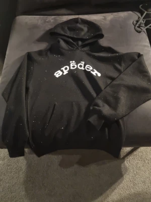Svart hoodie med glitter och tryck - Snygg svart hoodie med huva och glittriga detaljer över hela tyget. På bröstet står det 'spider' i vitt tryck med unik font. Perfekt för dig som vill sticka ut med en cool och trendig stil. Mjuk och bekväm, passar till de flesta outfits.