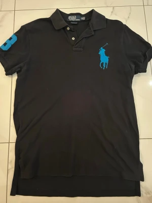Svart pikétröja från Ralph Lauren - Klassisk svart pikétröja från Ralph Lauren med blått broderat logotypmotiv på bröstet och siffran 3 på ärmen. Tröjan har krage, knappar framtill och är kortärmad. 