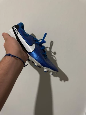 Blåa Nike fotbollsskor - Snygga blåa fotbollsskor från Nike med vit sula och svarta detaljer. Skorna har klassisk snörning och den ikoniska vita Nike-loggan på sidan. Dobbarna är utformade för bra grepp på planen. Perfekta för dig som vill sticka ut på matchen.