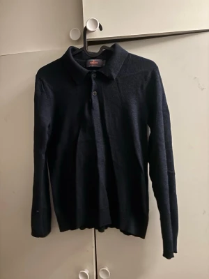 Mörkblå långärmad tröja med krage - En stilren mörkblå långärmad tröja med klassisk krage och knapp framtill. Tröjan har en enkel och clean design, perfekt för en chill och snygg look. Materialet ser ut att vara mjukt och bekvämt, passar bra till jeans eller chinos.