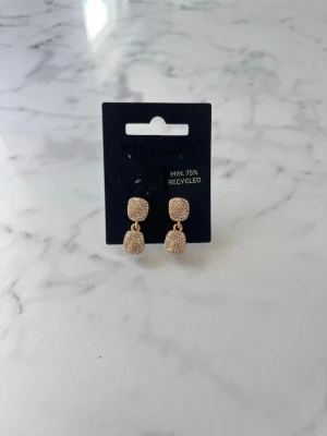 Guldiga glittriga örhängen - Säljer ett par snygga guldiga örhängen med dubbla fyrkantiga hängen täckta av små gnistrande stenar. Örhängena har en modern och trendig look och är tillverkade av minst 75% återvunnet material.