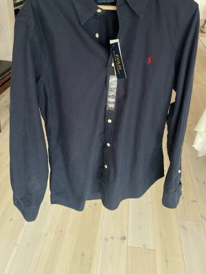 Mörkblå skjorta från Polo Ralph Lauren - Klassisk mörkblå skjorta från Polo Ralph Lauren med röd broderad logga på bröstet. Skjortan har långa ärmar, knappar framtill, en skjorta som kan användas både vinter och sommartid. Köpt i USA för 130 $ i RL butik men var för stor. Helt ny skick och en rejäl prissänkning mot vad jag köpte den för 