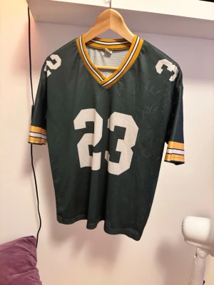 Grön Jersey t- shirt  -  grön Jersey med vita siffror 23 på bröstet och axlarna. T-shirten har gula och vita ränder på ärmarna och vid v-ringningen. Perfekt för en avslappnad och cool stil.