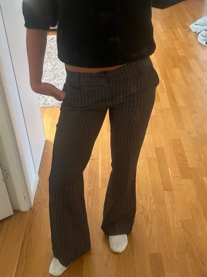 Gråa kostymbyxor med vita ränder - Säljer ett par gråa kostymbyxor med vita smala kritstreck. Byxorna har låg midja, vida ben och fickor fram. Klassisk dressad look med bälteshällor och knappstängning. Perfekta för dig som gillar en trendig och stilren vibe.