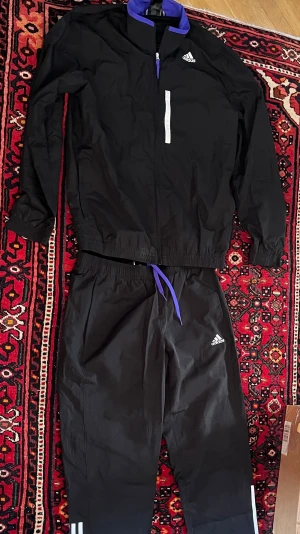 Svart Adidas träningsset med lila detaljer - Säljer ett svart träningsset från Adidas med jacka och byxor. Setet har vita Adidas-loggor, lila detaljer vid kragen och midjan samt vita ränder på byxbenen. Materialet är lätt och passar perfekt för träning eller uppvärmning.