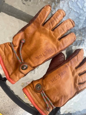 Bruna skinnhandskar från Hestra - Säljer ett par bruna skinnhandskar från Hestra med snygga detaljer och färgglad dragsko vid handleden. Handskarna har klassisk design med fem fingrar och är perfekta för kyliga dagar. Märket är präglat på ovansidan av båda handskarna. Nypris 1499kr