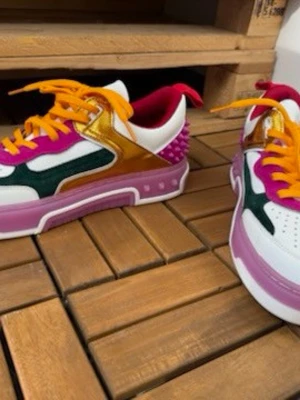 Färgglada sneakers med nitar och snörning - Unika sneakers i vitt, rosa, guld och grönt med lila detaljer och orange snören. Skorna har coola nitar bak och en chunky sula i rosa. Materialmix med inslag av skinn och syntet. Perfekta för dig som vill sticka ut med din stil.