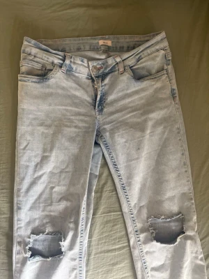 Ljusblå slitna jeans med hål - Säljer ett par ljusblå jeans med slitningar och stora hål vid knäna. Jeansen  har en cool detalj med rosa på bakfickan 