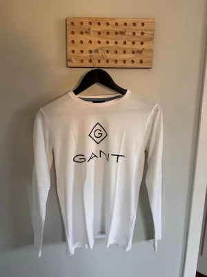 Vit långärmad t-shirt från GANT - Säljer en clean vit långärmad t-shirt från GANT med stor logga framtill. Tröjan har rund halsringning och är gjord i mjuk bomull, perfekt för en chill och stilren look.