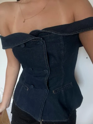 Mörkblå offshoulder korsettopp - Trendig mörkblå korsettopp i denim-look med offshoulder-design och markerad midja. Toppen har dekorativa knappar framtill och två fickdetaljer. Perfekt för dig som vill sticka ut med en edgy och unik stil.