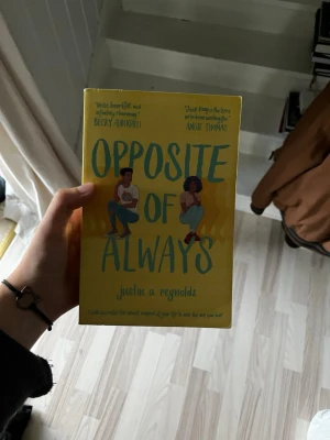 Opposite of Always av Justin A Reynolds - En ungdomsroman med gul mjukpärm och illustrationer av två personer på framsidan. Titeln 'Opposite of Always' är tryckt i stora blå bokstäver. Perfekt för dig som gillar romantik och tidsresor i modern miljö.
