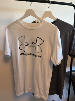 Under Armour t-shirt med logga - Säljer två snygga t-shirts från Under Armour, en i vitt och en i grått. Båda har stor logga framtill och klassisk rund hals. Perfekta till vardags eller träning, tillverkade i mjuk bomull för skön känsla. Enkel och stilren design som funkar till det mesta.