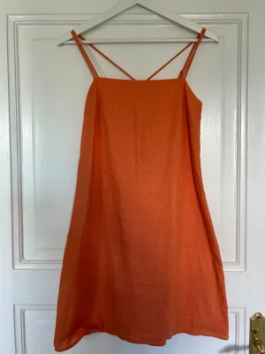 Orange ärmlös sommarklänning - Säljer en enkel och snygg orange klänning i linne med smala axelband och rak passform. Perfekt för varma dagar och ger en avslappnad vibe. Klänningen är kort och har en minimalistisk design utan mönster eller extra detaljer.