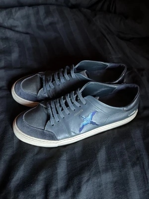 Svarta Arigatos - Snygga svarta sneakers i läder med blå broderad fågel på sidan och vita sulor. Skorna har svarta skosnören och detaljer i mocka vid framsidan. 