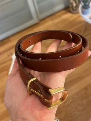 Brunt skärp i skinn med guldfärgat spänne - Snyggt brunt skärp i äkta skinn med stilrent guldfärgat metallspänne. Skärpet är tillverkat i Sverige och har en klassisk design som passar till många olika outfits. Perfekt accessoar för att lyfta din look.