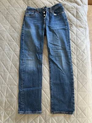 Blå raka jeans från Levi's - Klassiska blå jeans från Levi's med rak passform och knappgylf. Jeansen har fem fickor, kontrastsömmar och den ikoniska läderlappen bak i midjan. Tillverkade i slitstark denim som ger en tidlös look.