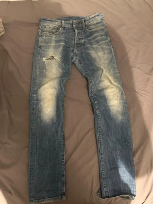 G star raw blåa jeans  - Ett par blå jeans med slim fit och slitningar, inklusive ett hål på vänster lår. Jeansen har klassisk femficksdesign och ljusa tvättade partier framtill och baktill. Materialet är denim i bomull. Perfekta för en avslappnad och trendig stil.