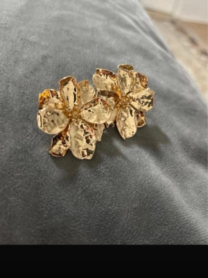 Guldiga blomformade örhängen - Säljer ett par statement örhängen i form av blommor med hamrad yta. Örhängena är i guldton och har en cool, unik design som verkligen sticker ut. Perfekta för dig som vill addera något extra till din look!