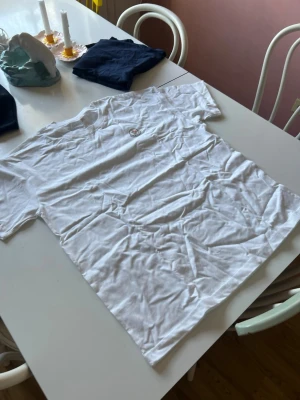 Vit Moncler t-shirt i bomull - Säljer en klassisk vit t-shirt från Moncler med rund hals och det ikoniska Moncler-logotypen på bröstet. T-shirten är i mjuk bomull och har en enkel, clean look som passar till allt. Är XL men sitter som L