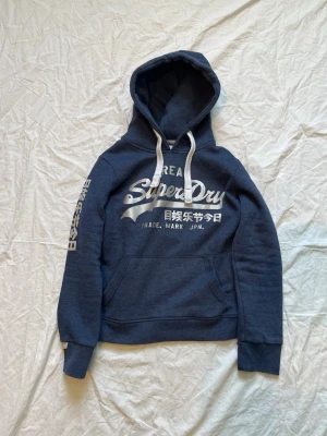 Mörkblå hoodie från Superdry - Mörkblå hoodie från Superdry med vit logga och japansk text på bröstet samt tryck på ena ärmen. Klassisk känguruficka framtill och vita dragsnören i huvan. Tillverkad i mjukt bomullsmaterial för extra komfort.