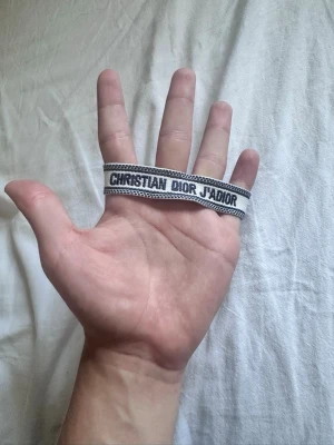 Christian Dior J'Adior tygarmband - Snyggt tygarmband från Christian Dior med texten 'J'Adior' i svart på vit bakgrund. Armbandet har flätade band och tofsar i ändarna, vilket ger en cool och trendig look. Perfekt accessoar för att lyfta din outfit med en exklusiv touch.