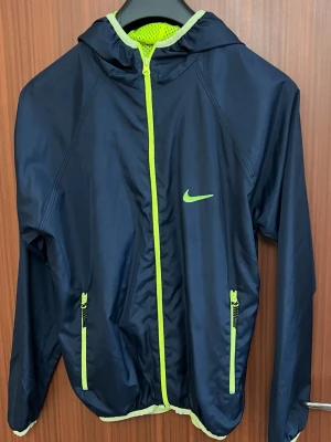 Marinblå Nike vindjacka med huva - Snygg marinblå vindjacka från Nike med huva och dragkedja i neonfärg. Jackan har två fickor med dragkedja och meshfoder i neon för extra ventilation. Perfekt för löpning eller utomhusaktiviteter. Nike-logga på bröstet och sportig design.