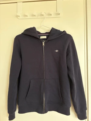 Mörkblå hoodie med dragkedja från Gant - Mörkblå hoodie från Gant med dragkedja framtill och två stora fickor. Liten broderad Gant-logga på bröstet. Storlek 158/164. Är i mycket bra skick