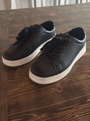 Svarta sneakers med vit sula - Stilrena svarta sneakers med tjock vit sula och svarta skosnören. Skorna har en klassisk låg profil och är tillverkade i ett slätt, läderliknande material. Perfekta för en clean och enkel look.