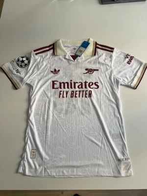Helt ny! Arsenal 25/26 tredje Eze - Säljer en vit Arsenal matchtröja från Adidas med vinröda detaljer och klubbmärke. Tröjan har trycket 'Emirates Fly Better' på bröstet, Champions League-märke på ärmen och nummer 10 med namnet EZE på ryggen. Klassisk krage och korta ärmar.