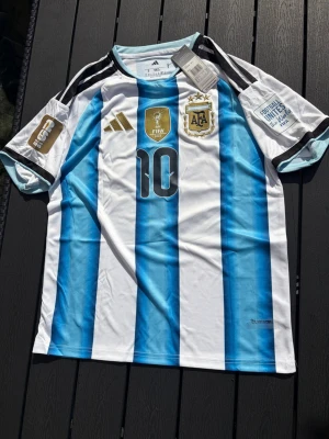 Argentina Messi matchtröja blå/vit - Säljer en officiell Argentina fotbollströja från Adidas med Messi och nummer 10. Tröjan har blå och vita vertikala ränder, svarta detaljer och guldiga loggor. Materialet är lätt och andas, perfekt för fotbollsträning eller match.