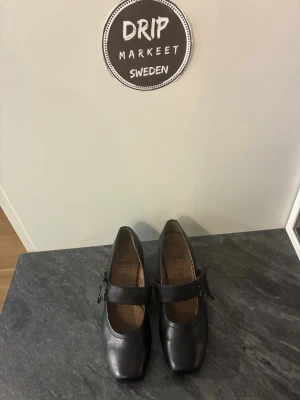  Caprice Svarta ballerinaskor med rem - Stilrena svarta ballerinaskor med bred rem över foten och fyrkantig tå. Skorna har en klassisk design och är tillverkade i ett material som liknar läder. Perfekta för dig som gillar en enkel men trendig look.