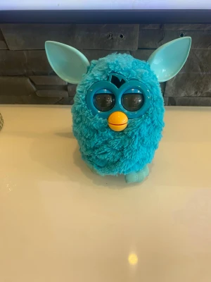 Blå Furby-leksak med stora öron - Säljer en blå Furby-leksak med fluffig päls, stora plastöron och en orange näbb. Den har stora ögon med plast runt och en liten blå svans baktill. Perfekt för dig som gillar samlarprylar eller vill ha en rolig prydnad.