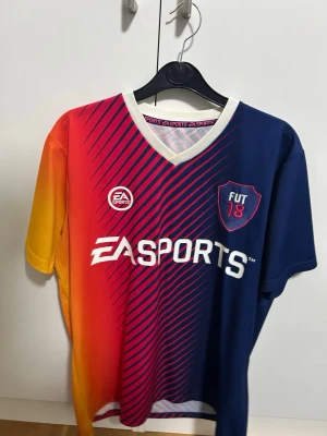 EA Sports FUT 18 fotbollströja storlek M - Säljer en färgstark EA Sports FUT 18 fotbollströja med gradient i blått, rött och gult. Tröjan har vita detaljer, V-ringad krage och tryckta loggor framtill samt stort nummer 18 på ryggen. Perfekt för dig som gillar gaming och sport.