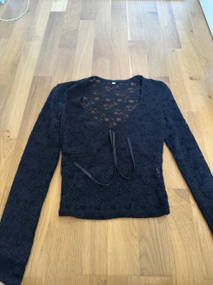 Svart spetsblus med knytband - Säljer en svart långärmad blus i spets med v-ringning och knytband framtill. Blusen är genomskinlig och har ett snyggt blommigt spetsmönster som ger en cool och elegant look. Perfekt att styla med jeans eller kjol för en trendig outfit.