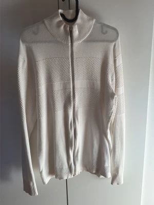Vit stickad ziptröja från Selected Homme - Vit stickad tröja från Selected Homme med hel dragkedja framtill och diskret strukturmönster över bröstet och ärmarna. Hög krage och ribbade muddar ger en clean och stilren look. Perfekt för lager-på-lager under kyliga dagar.