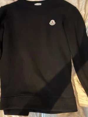 Svart sweatshirt från Moncler - Svart sweatshirt från Moncler med rund halsringning och Moncler-logga på bröstet. Tröjan har en stilren design och är tillverkad i mjukt material, perfekt för en avslappnad och trendig look.