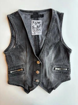 Svart jeansväst med dragkedjor storlek 134/140/ XXS - Snygg svart jeansvästt i tvättad denimlook med djup V-ringning, tre metallknappar framtill och två fickor med dragkedjor. Västen har justerbart spänne i ryggen för perfekt passform. Storlek 134/140/ XXS