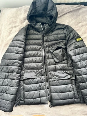 Svart dunjacka från Barbour L - Snygg svart dunjacka från Barbour International med huva och dragkedja. Jackan har tre stora fickor framtill med lock och tryckknappar samt Barbour-märke på ärmen. Tillverkad i lätt och värmande fiberdun, perfekt för kalla vinterdagar. I storlek L men passar även M.