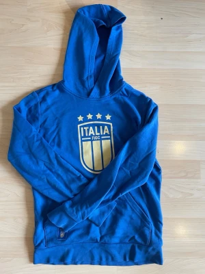 Blå adidas Italia hoodie  - Snygg blå hoodie med stort gult Italia-tryck och fyra stjärnor på bröstet. Hoodien har huva och magficka och långa ärmar. Perfekt för dig som gillar fotboll och vill representera Italien med stil. Storlek: Uk: 13-14 years   Material:polyester,bomull