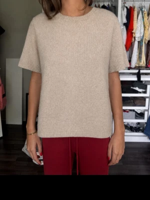 Beige stickad t-shirt från Soft Goat - Säljer en beige stickad t-shirt från Soft Goat i mjuk och lyxig kvalitet. Tröjan har korta ärmar, rund halsringning och ribbade kanter. Perfekt för dig som gillar minimalistisk och stilren design. Materialet känns riktigt skönt mot huden.