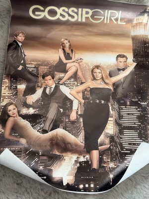 Gossip Girl poster - Snygg poster med motiv från tv-serien Gossip Girl. Affischen har en stadssilhuett i bakgrunden och visar huvudkaraktärerna i eleganta kläder. Färgerna går i svart, beige och guldtoner. Perfekt för dig som gillar serier och vill ha cool väggdekor.