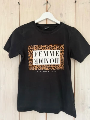 Svart t-shirt med leopardmönster Gina Tricot - Cool svart t-shirt från Gina Tricot med tryck framtill där det står 'FEMME HOMME' och 'NEW YORK CITY' i vitt. Runt texten är det en ruta med leopardmönster. Klassisk rund hals och korta ärmar. Tillverkad i mjuk bomull.