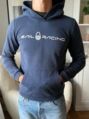 Mörkblå hoodie från Sail Racing - Mörkblå hoodie från Sail Racing med vit logga och text på bröstet. Klassisk känguruficka framtill och huva. Tillverkad i mjukt bomullsmaterial och har ribbade muddar vid ärmslut och nederkant. Perfekt för en avslappnad och sportig stil. M I BARN!!!