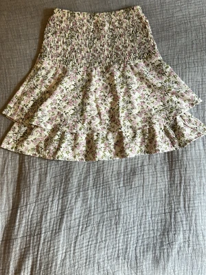 Blommig volangkjol med smock - Supersöt kjol med smock i midjan och dubbla volanger. Tyg i vitt med små rosa och gröna blommor. Motsvarar storlek 134-140