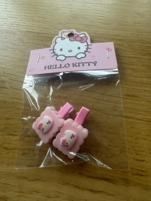 Hello Kitty rosa hårspännen - Säljer ett par söta rosa hårspännen med Hello Kitty-motiv. Spännena har en fluffig kant och dekoreras med en liten rosett. Perfekta för att piffa upp frisyren med en gullig touch. Materialet är plast och metall.