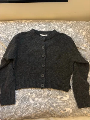 Mörkgrå cropped kofta med knappar - Mysig mörkgrå cropped kofta med rund hals och stora matchande knappar framtill. Koftan har långa ärmar och ribbade muddar vid ärmslut och nederkant. Perfekt att slänga över en t-shirt för en chill look.
