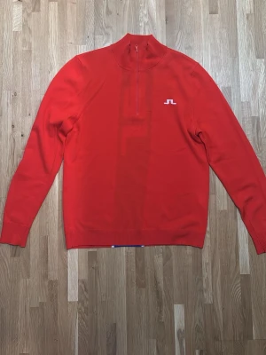 Röd stickad J.Lindeberg tröja med blå detalj - Röd stickad tröja med halv dragkedja och vit logga på bröstet. Baksidan har en bred blå rand med vit logga i mitten. Långa ärmar och ribbade muddar. Perfekt för dig som gillar färgstarka plagg och sportig stil.